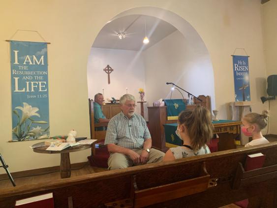 Decorative image of pastor conducting a childens sermon, taken from a pew near the front of the church and looking forward.